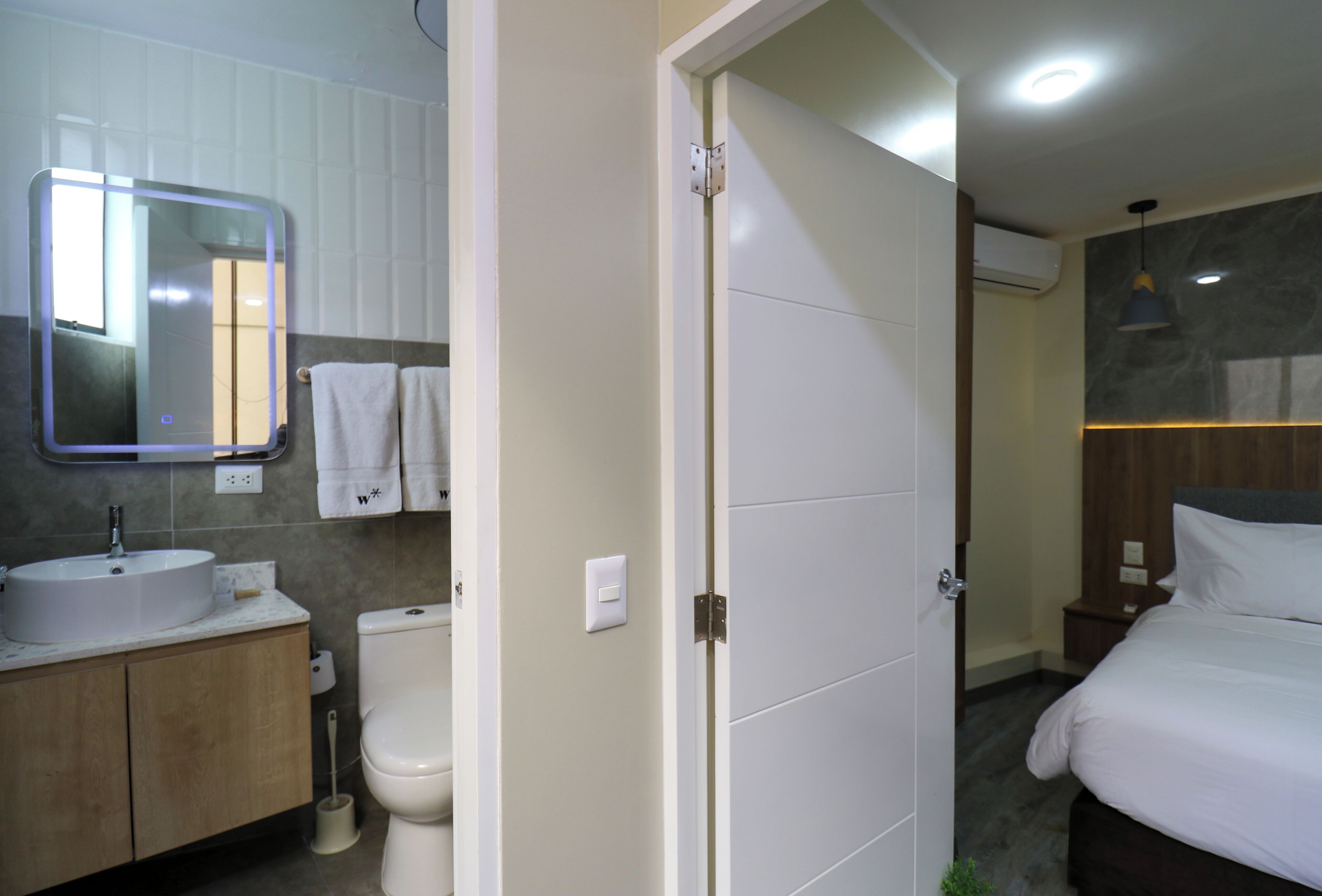 Une salle de bain moderne avec un lavabo, des toilettes et un miroir. Une porte donne sur une chambre à coucher équipée d’un lit et d’une table de chevet.