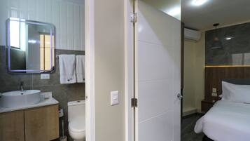 Une salle de bain moderne avec un lavabo, des toilettes et un miroir. Une porte donne sur une chambre à coucher équipée d’un lit et d’une table de chevet.