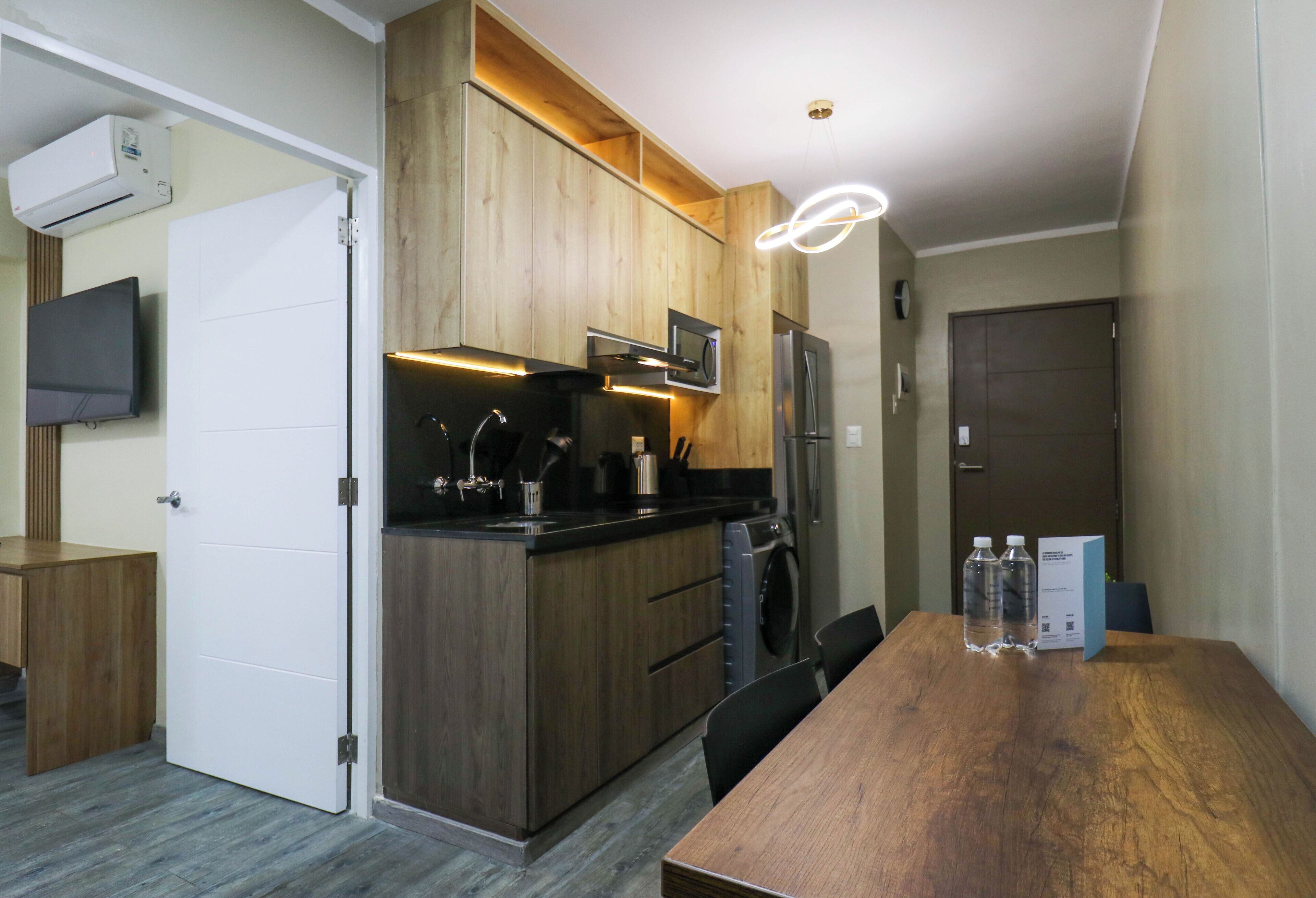 A modern kitchen with wooden cabinets, a microwave, a refrigerator, and a built-in oven. There is a dining table with chairs and two water bottles on it.
