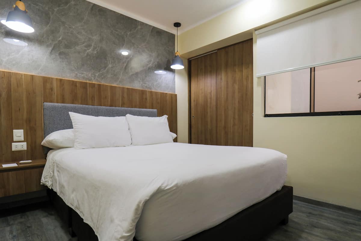 A modern hotel room with a large bed, wooden paneling, a marble accent wall, and a window with blinds.