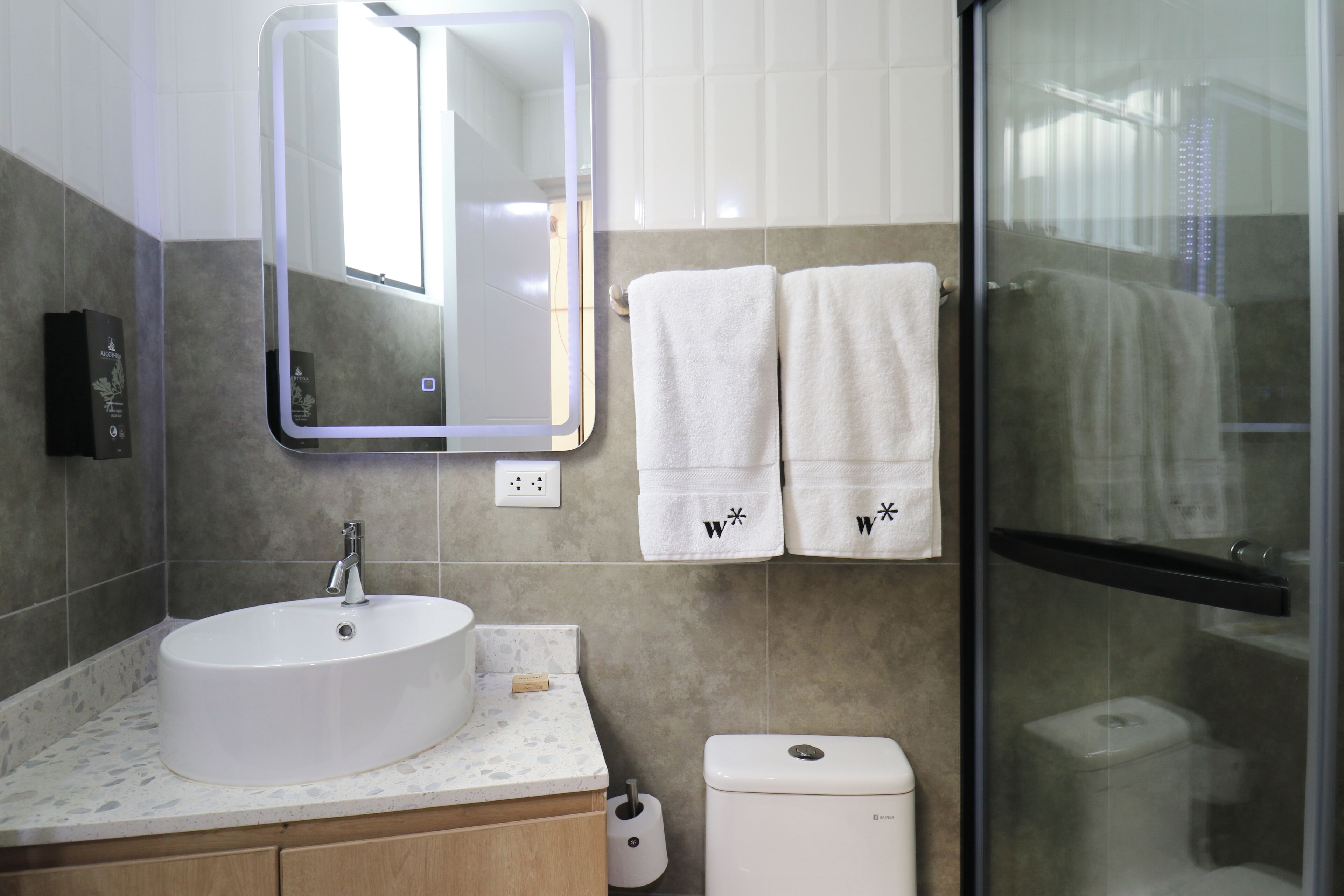 A bathroom with a round sink, a mirror, two towels with a logo, a toilet, and a glass shower enclosure.
