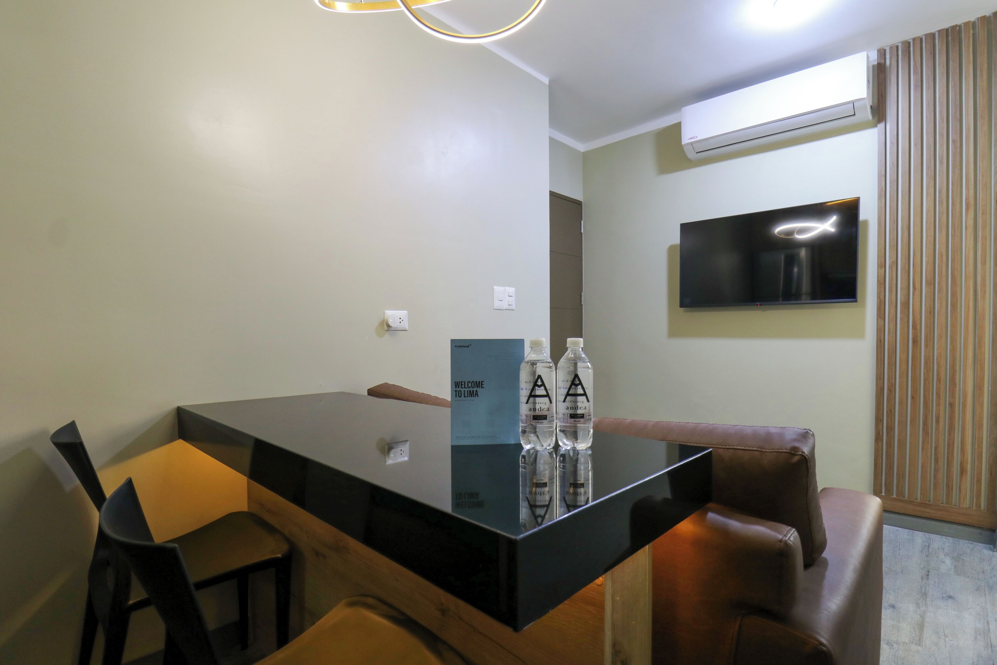 A modern dining area with a black glass table, two chairs, and a brown sofa. A flat-screen TV is mounted on the wall, and there are two bottles on the table.
