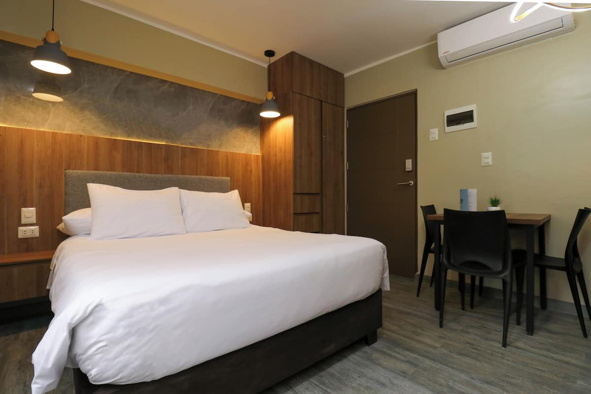 A hotel room with a large bed, wooden paneling, a desk with chairs, and an air conditioning unit.