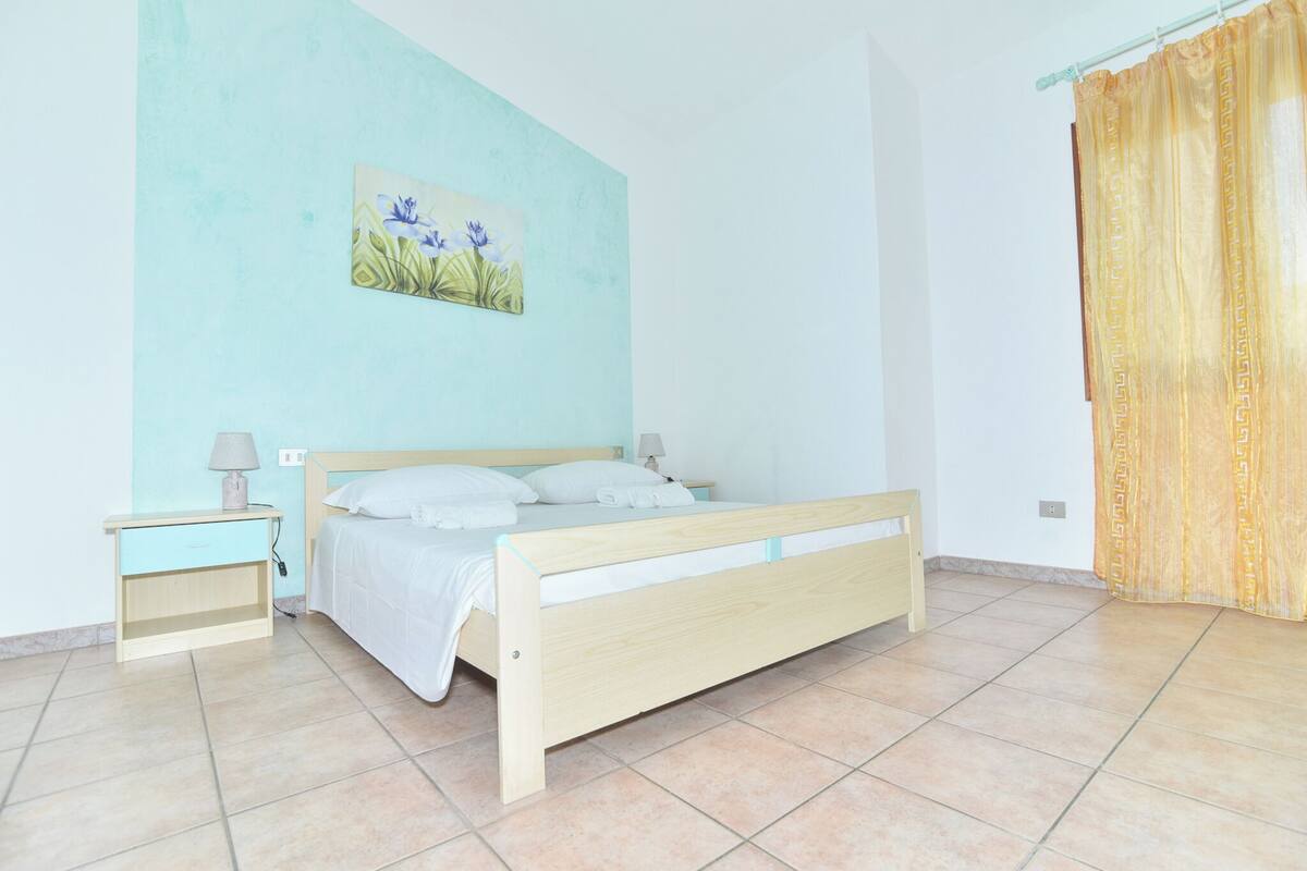 A bedroom with a bed, nightstands, a wall-mounted painting, and a curtain.