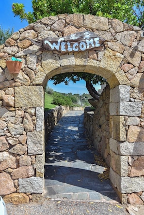 A stone archway with a 'Welcome' sign, leading to a stone pathway. - La Costa View (Arzachena)