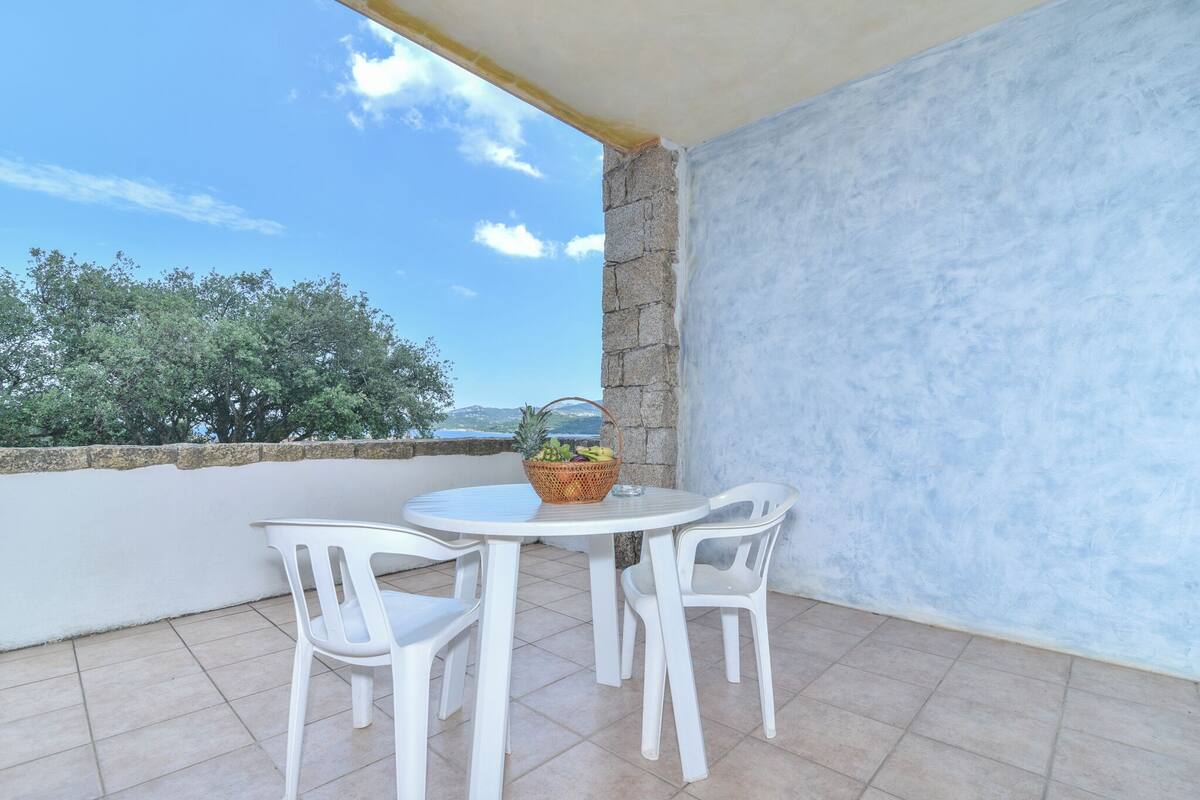 A balcony with a white table and chairs, a wicker basket with fruits, and a view of trees and the sea.