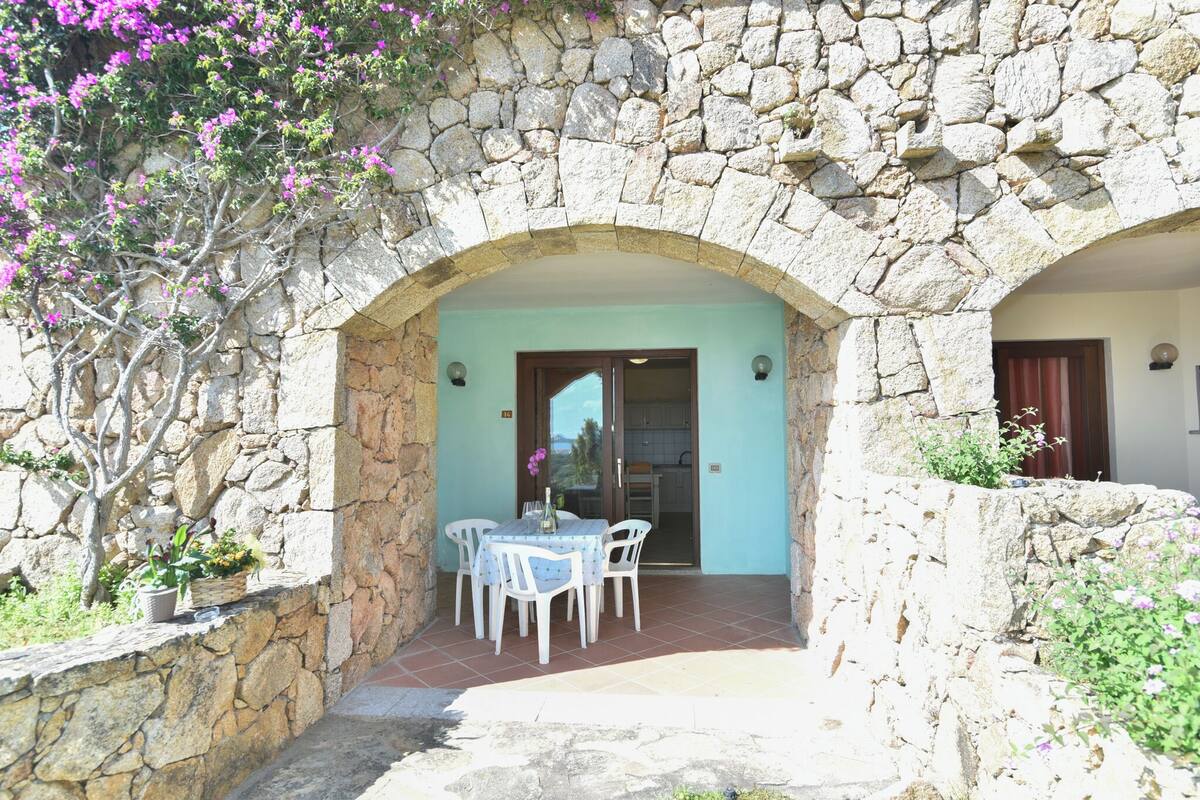 A stone archway leading to a covered patio with a table and chairs, surrounded by potted plants and a stone wall.