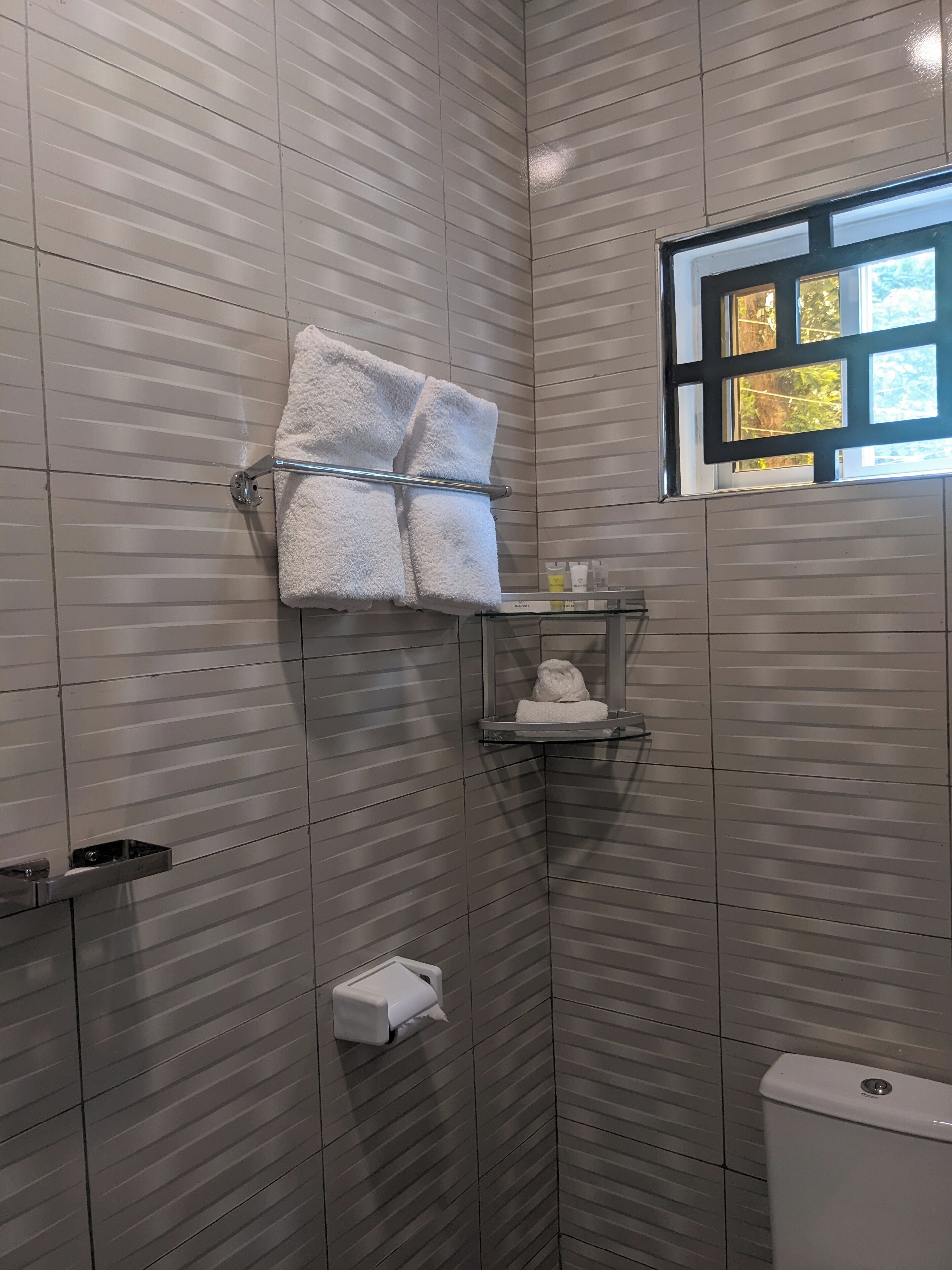 Une salle de bain moderne avec des murs recouverts de carreaux à rayures, une étagère en verre sur laquelle repose une serviette, un porte-rouleau de papier toilette et une fenêtre donnant sur des arbres.