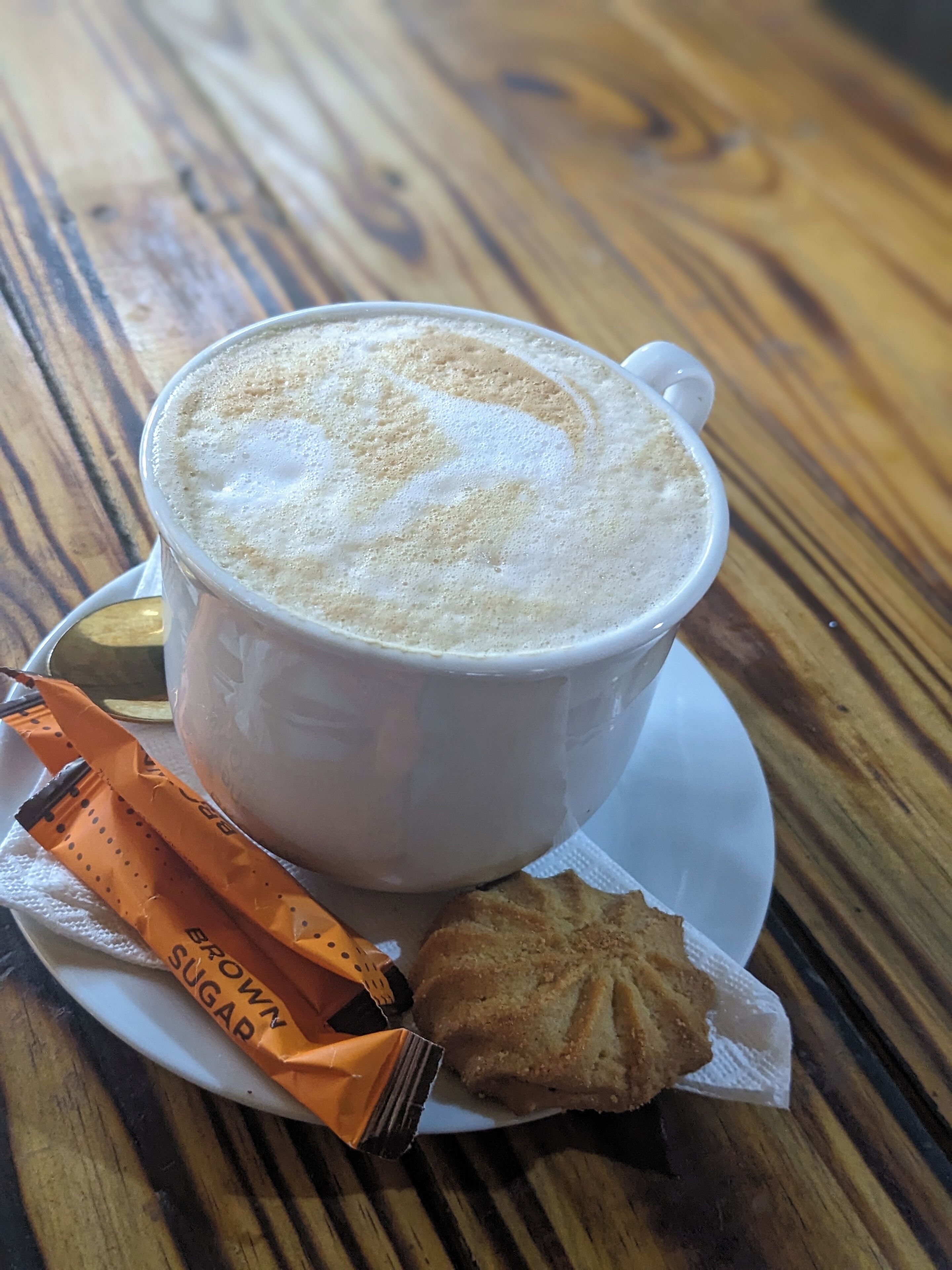 A cup of coffee with foam on top, a biscuit, and a packet of sugar on a wooden table.