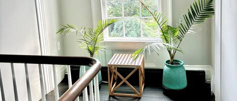 Un escalier avec une rampe blanche mène à un palier. Sur le palier, on trouve une petite table en bois surmontée d’un plateau en métal ajouré, entourée de plantes en pot. Une grande fenêtre donnant sur un paysage verdoyant est visible à l’arrière-plan.