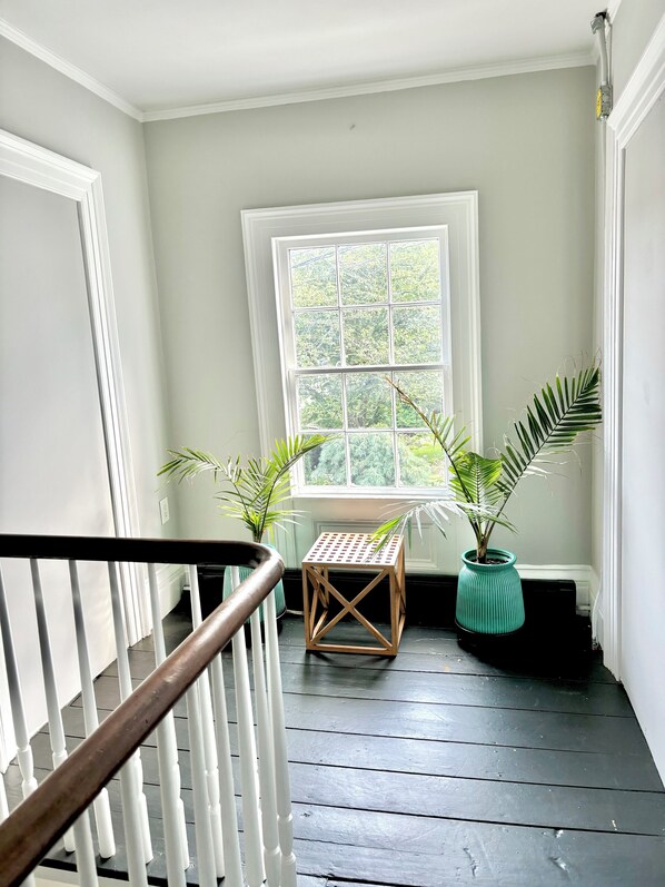 A staircase with a white railing leading to a landing. There is a small wooden table with a metal grid top on the landing, surrounded by potted plants. A large window with a view of greenery is visible in the background.