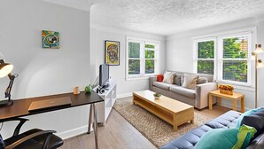 A modern living room with a sofa, coffee table, and a desk with a lamp. There are two windows with blinds, and artwork on the walls.
