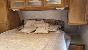 A bedroom with a bed, a wooden headboard, a bedside table, and a small drawer. - The Riverside Fleetwood.   (Petit Étang)
