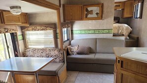 A compact RV interior with a kitchenette, a sofa, a dining table, and a small bed. - The Riverside Fleetwood.   (Petit Étang)