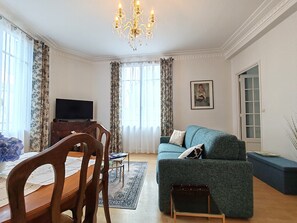 A living room with a wooden dining table, chairs, a sofa, a TV, and a chandelier. - Appartement Le Baron Cœur de Rouen Avec Petit Déjeuner Inclus - Normandie (Rouen)