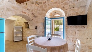 A stone-walled room with a round table, white chairs, a flat-screen TV, and a view through a glass door.