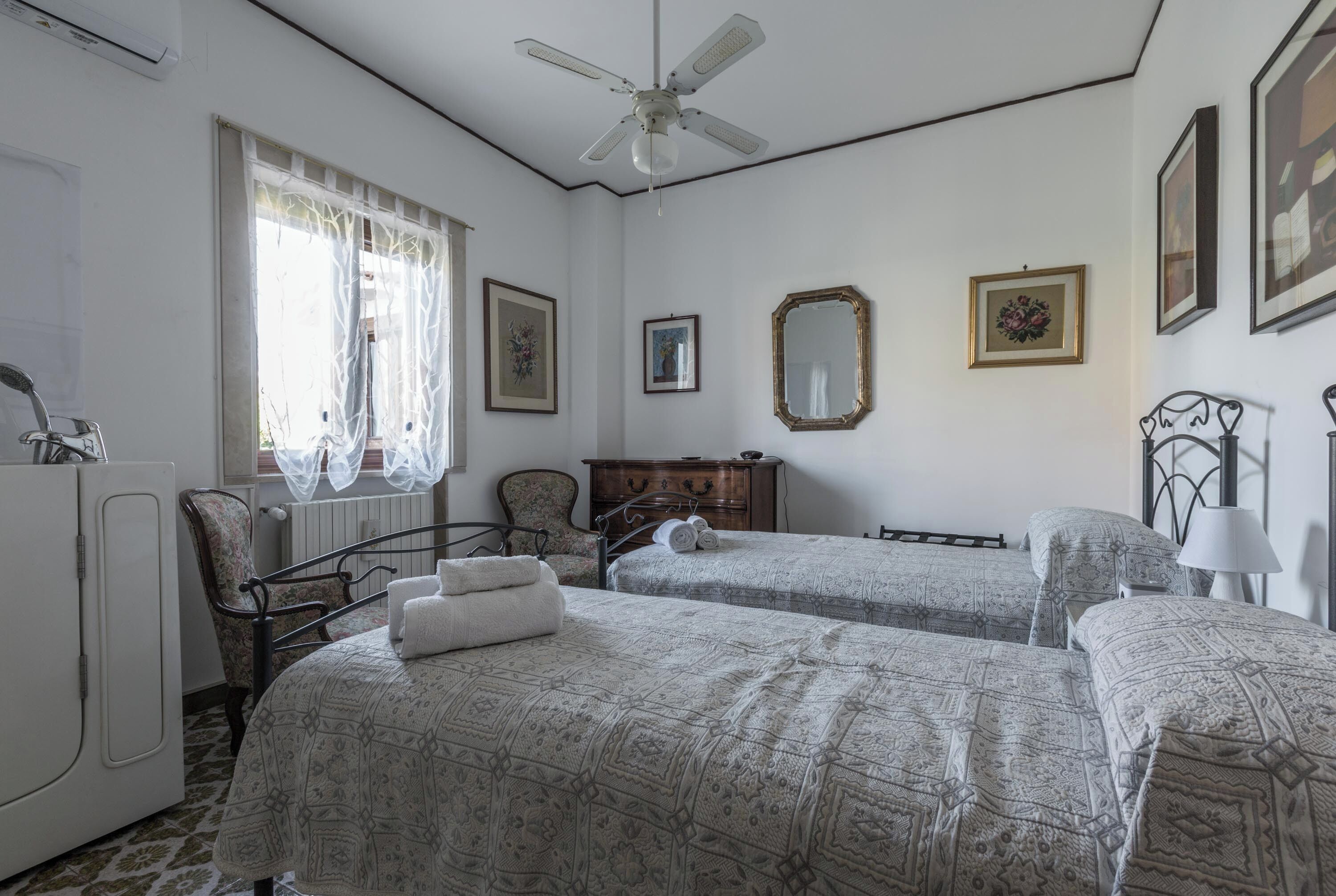 Ein Schlafzimmer mit zwei Betten, einem Heizkörper, einem Sessel, einer Kommode und einem Deckenventilator.