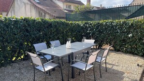 Patio esterno con tavolo rettangolare e sei sedie, circondato da ghiaia e siepe.