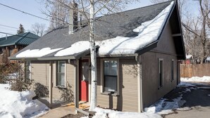 Una pequeña casa de un piso con techo inclinado cubierto de nieve, una puerta roja y un jardín delantero visible con acumulación de nieve.