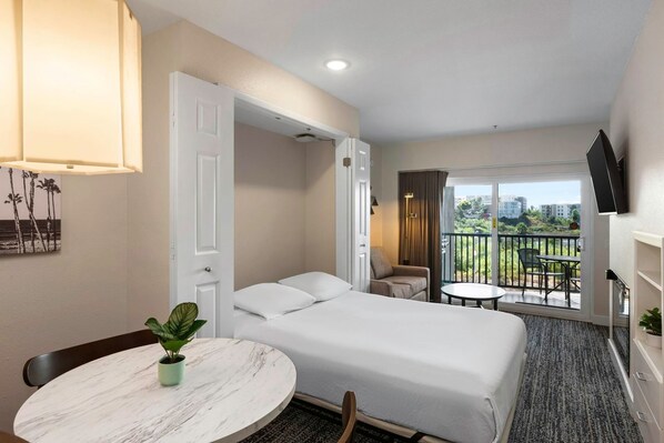 Una habitación de hotel con una cama, una mesa redonda con una planta, un balcón con una silla y un televisor instalado en la pared.