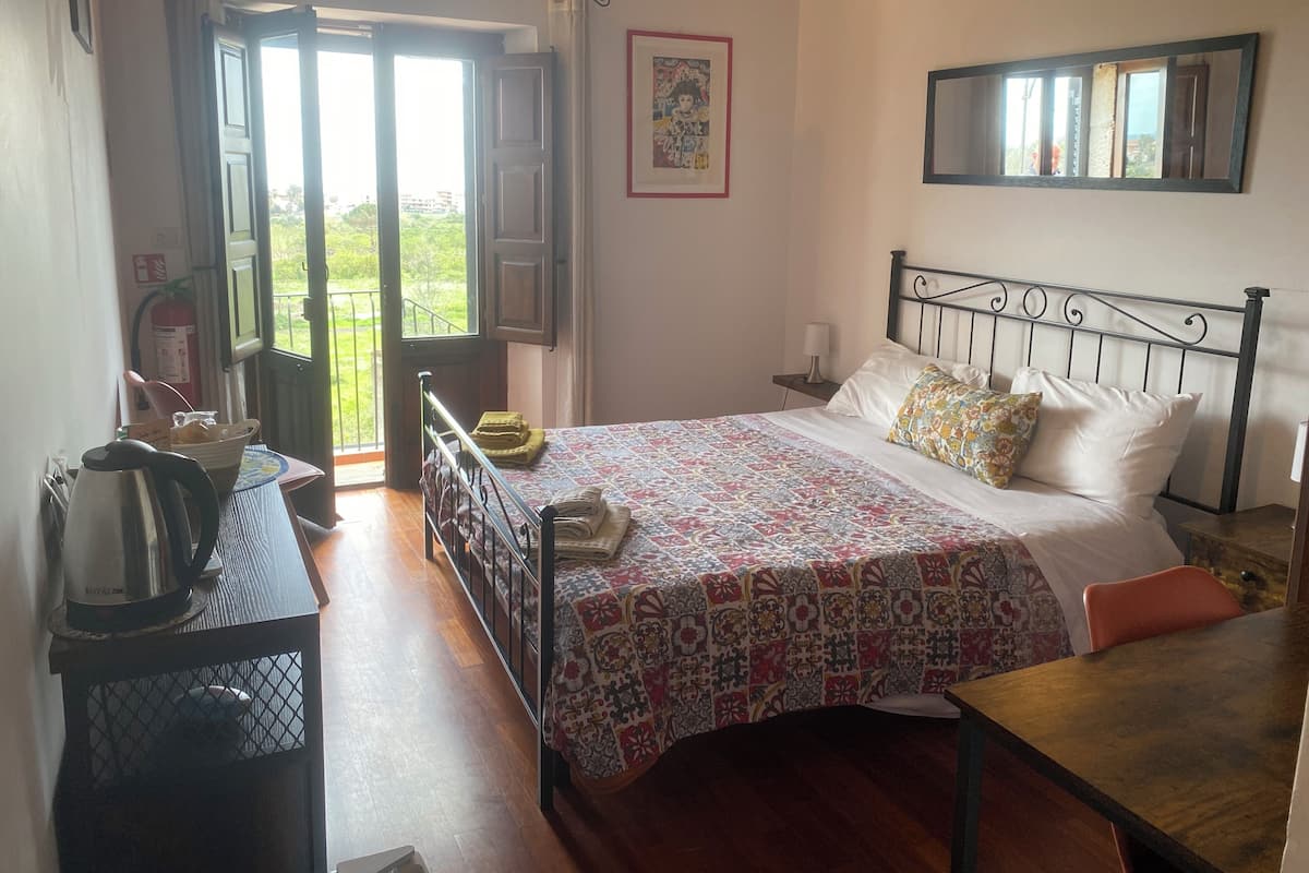 Un dormitorio con cama, una mesa, una silla, un ventanal con cortinas, un aire acondicionado instalado en la pared y un cuadro enmarcado.
