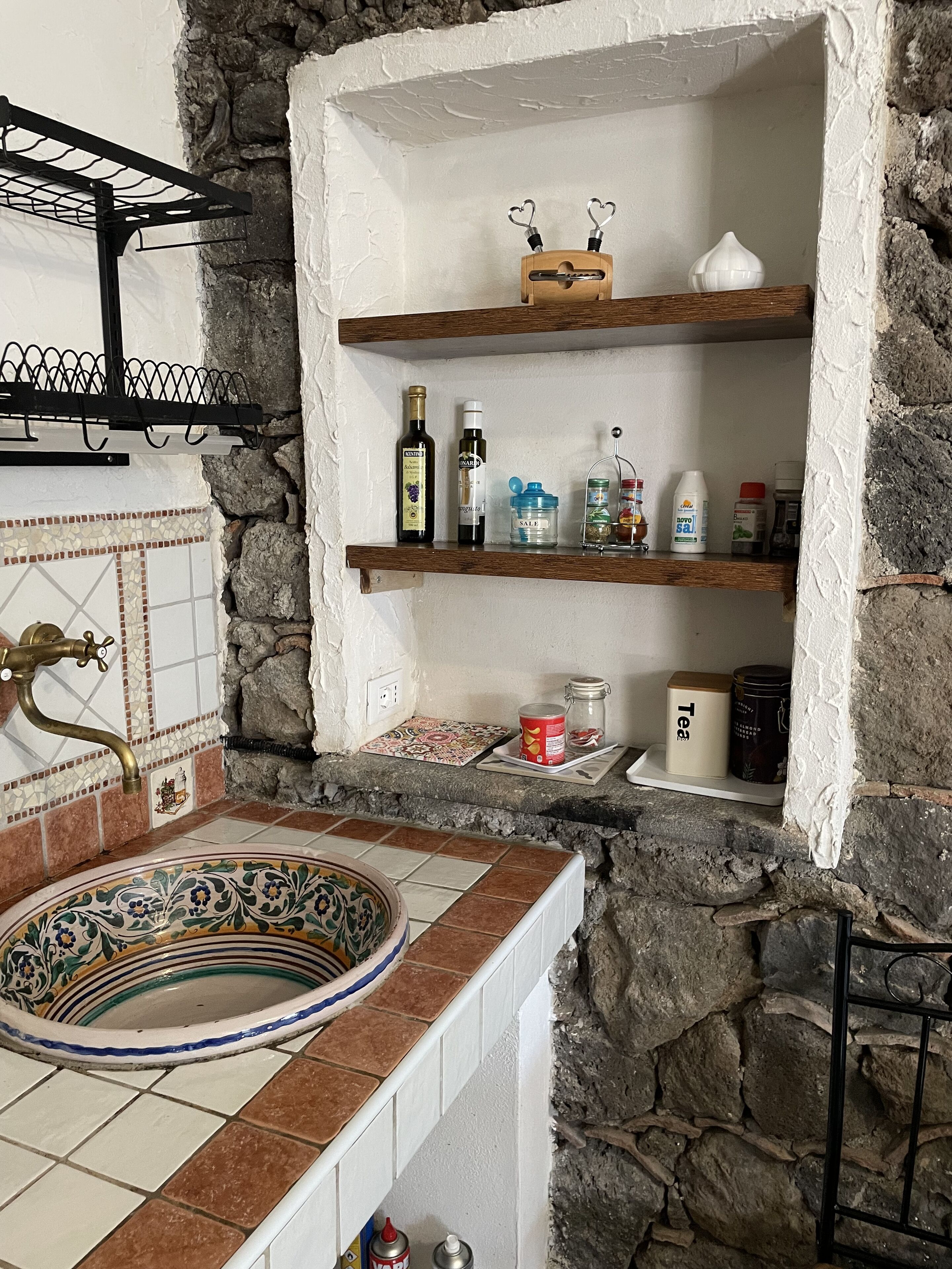 Un baño con pared de piedra, lavabo con azulejos, repisas con artículos de aseo y un perchero.