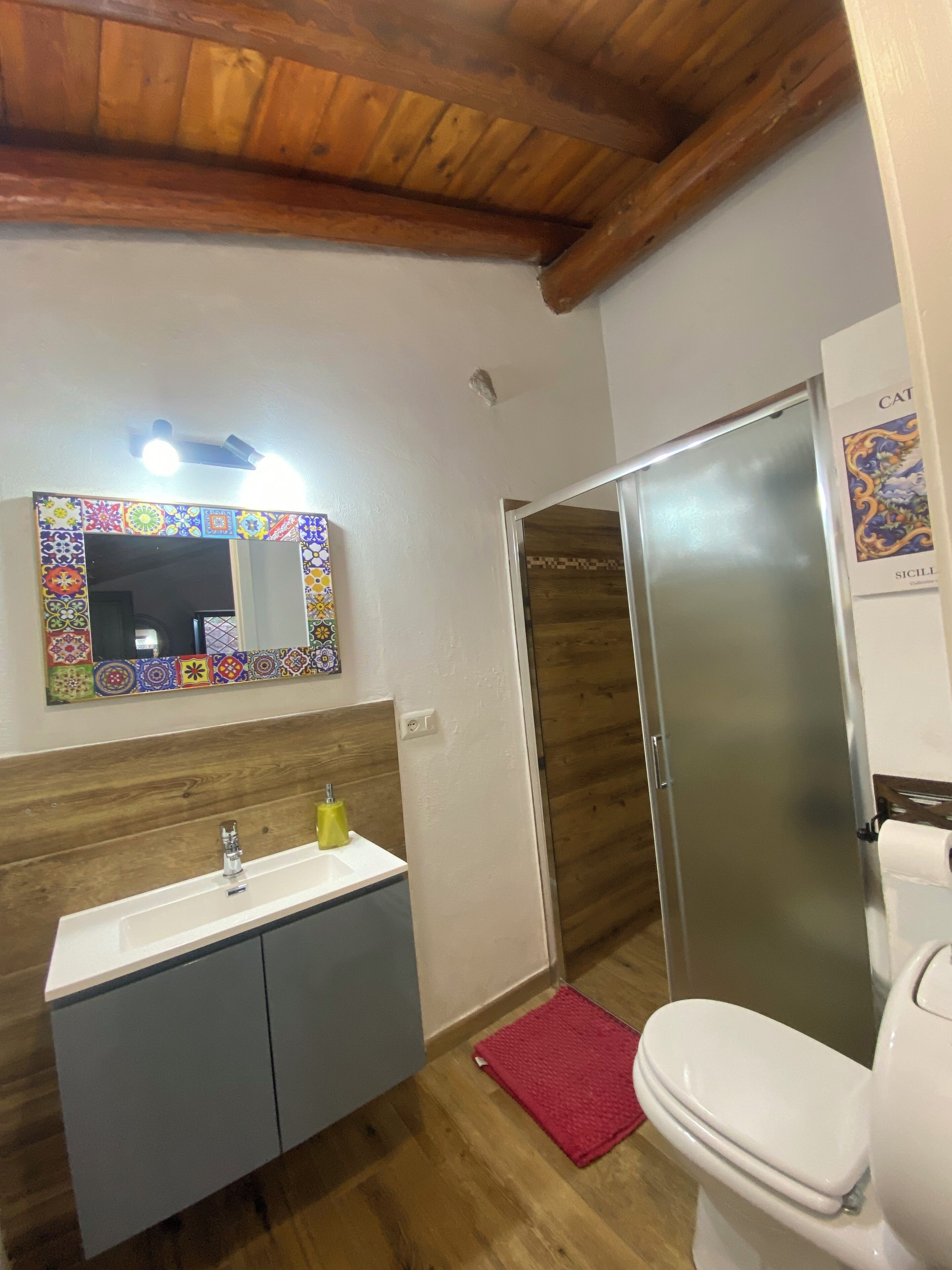 Un baño con techo de madera, lavamanos con espejo, inodoro y puerta de ducha.