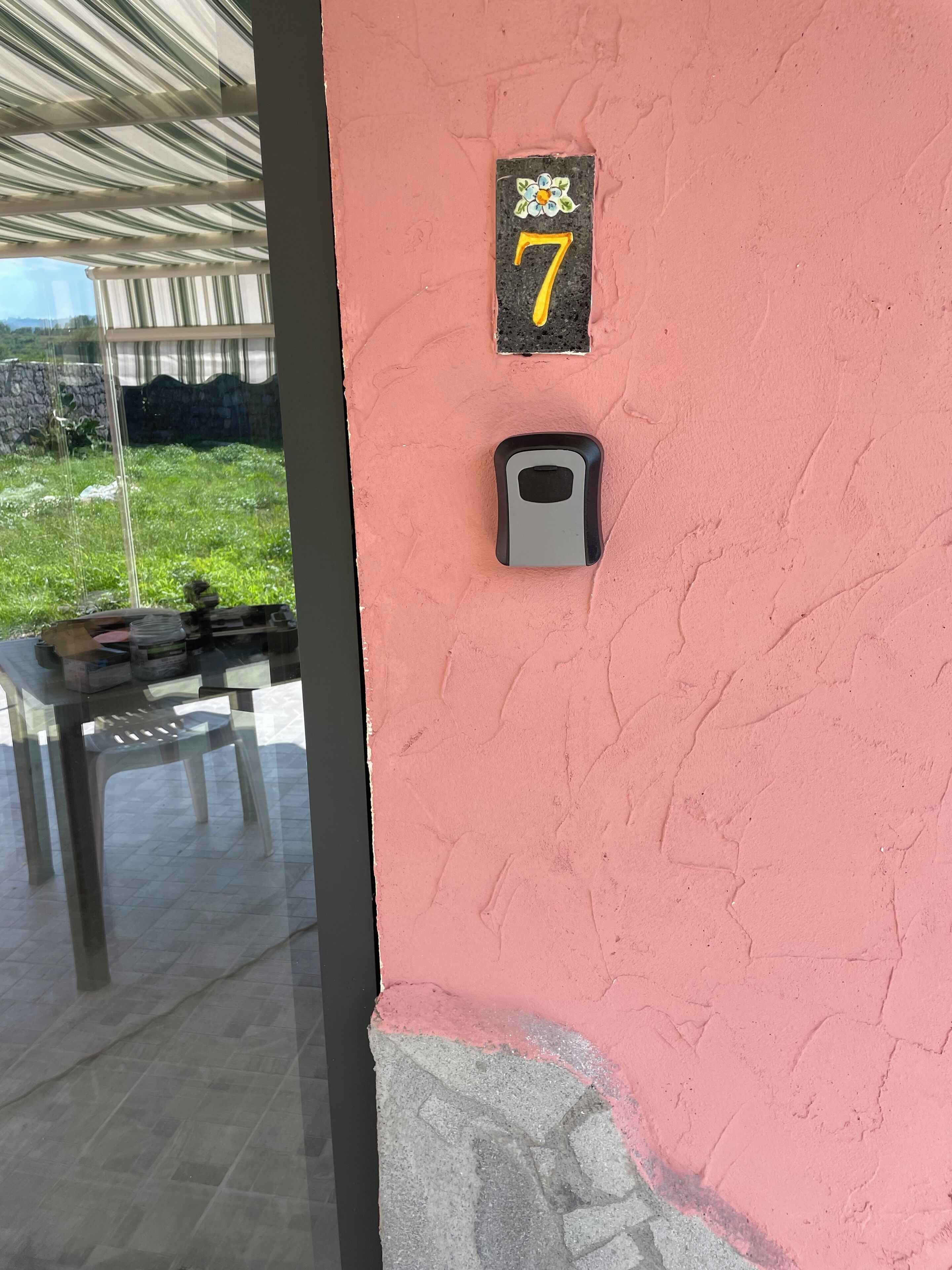 Un muro rosa con un cartello con il numero '7', un campanello e una porta in vetro che si apre su uno spazio esterno con un tavolo e sedie.