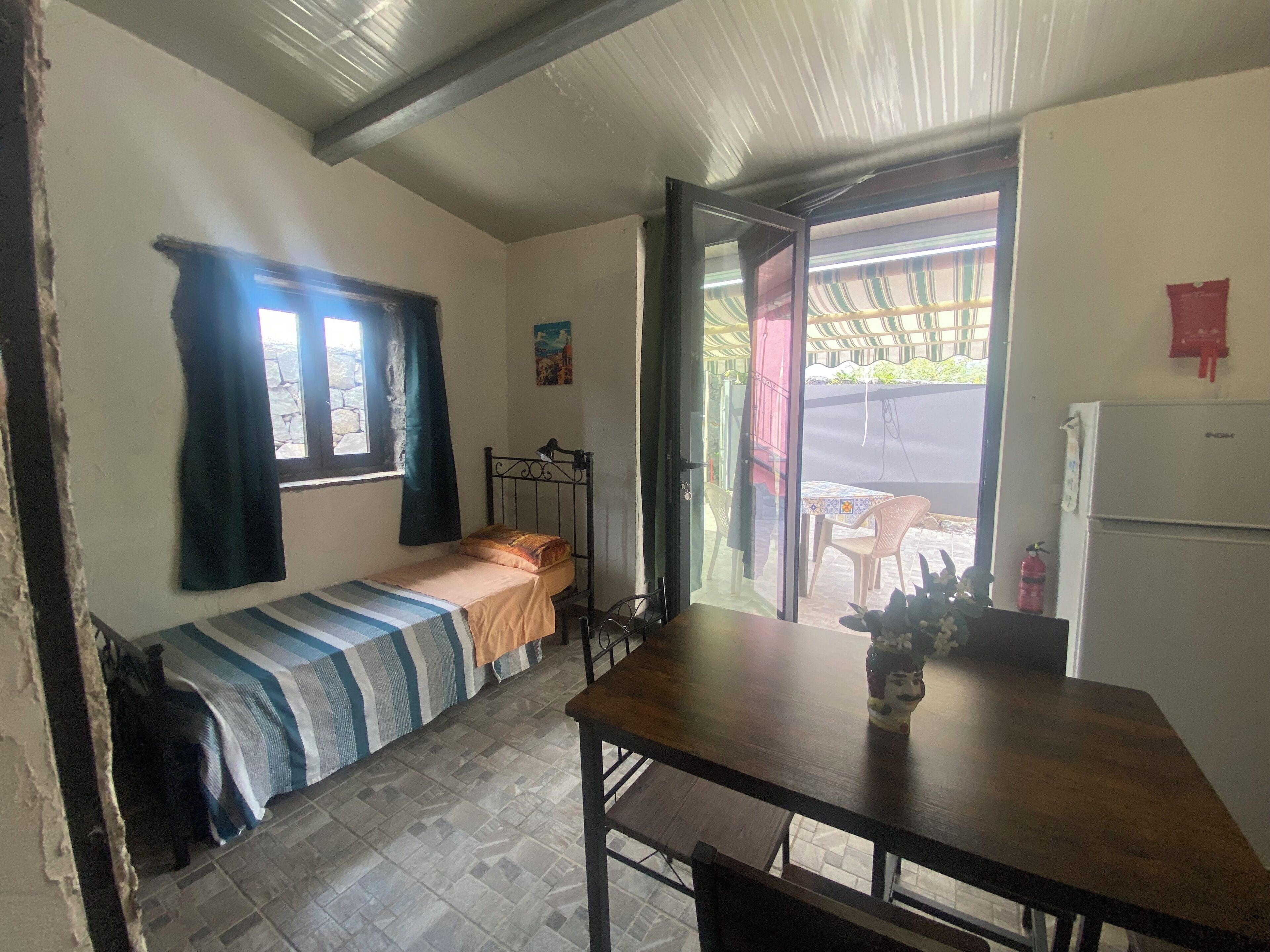 Una habitación pequeña y funcional con una cama individual, una mesa y una silla. Cuenta con una ventana con cortinas, una puerta que da a un espacio exterior y un refrigerador.