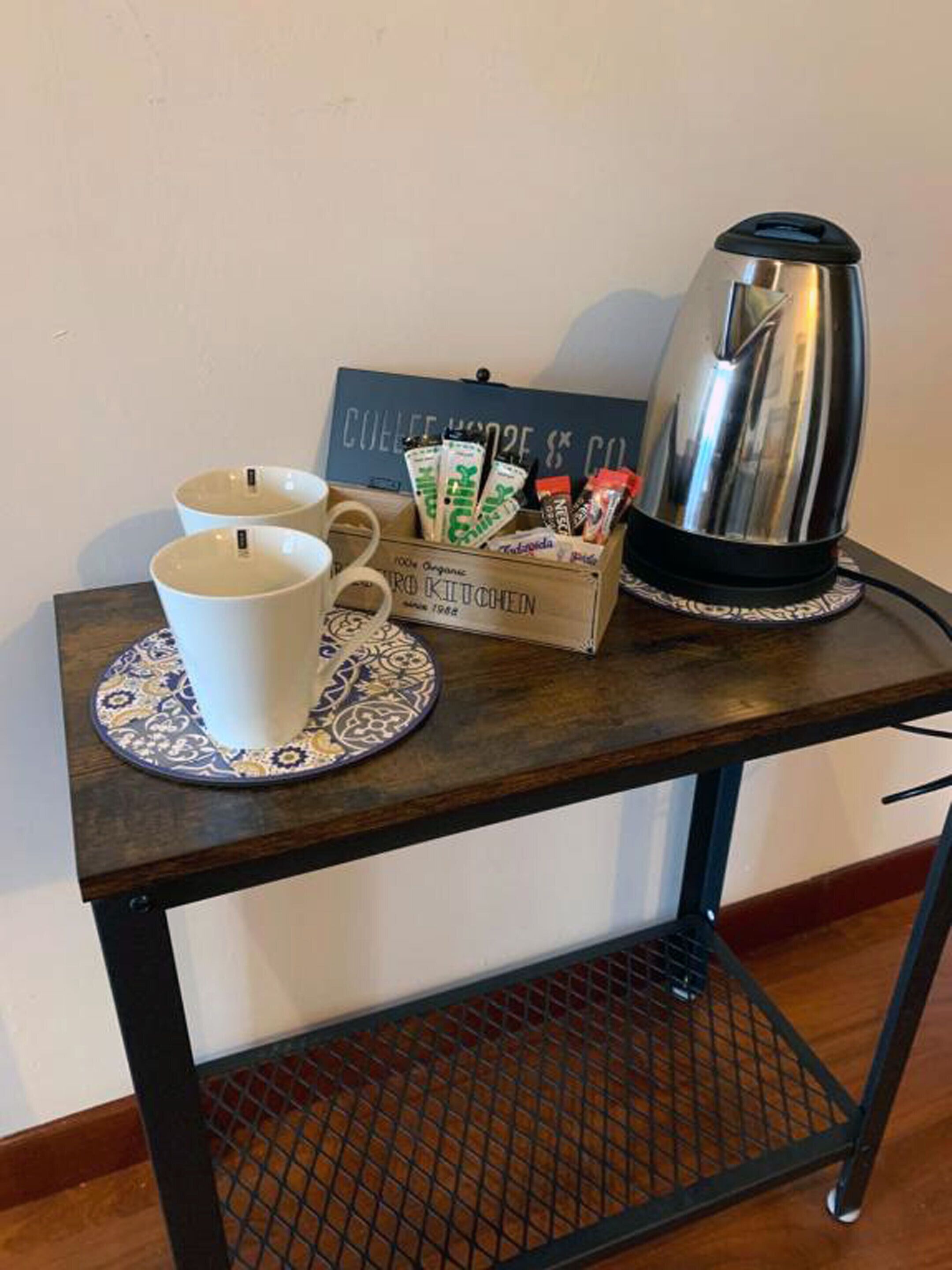 Un coin café avec une bouilloire, deux tasses et une boîte de capsules de café, le tout posé sur une table en bois.
