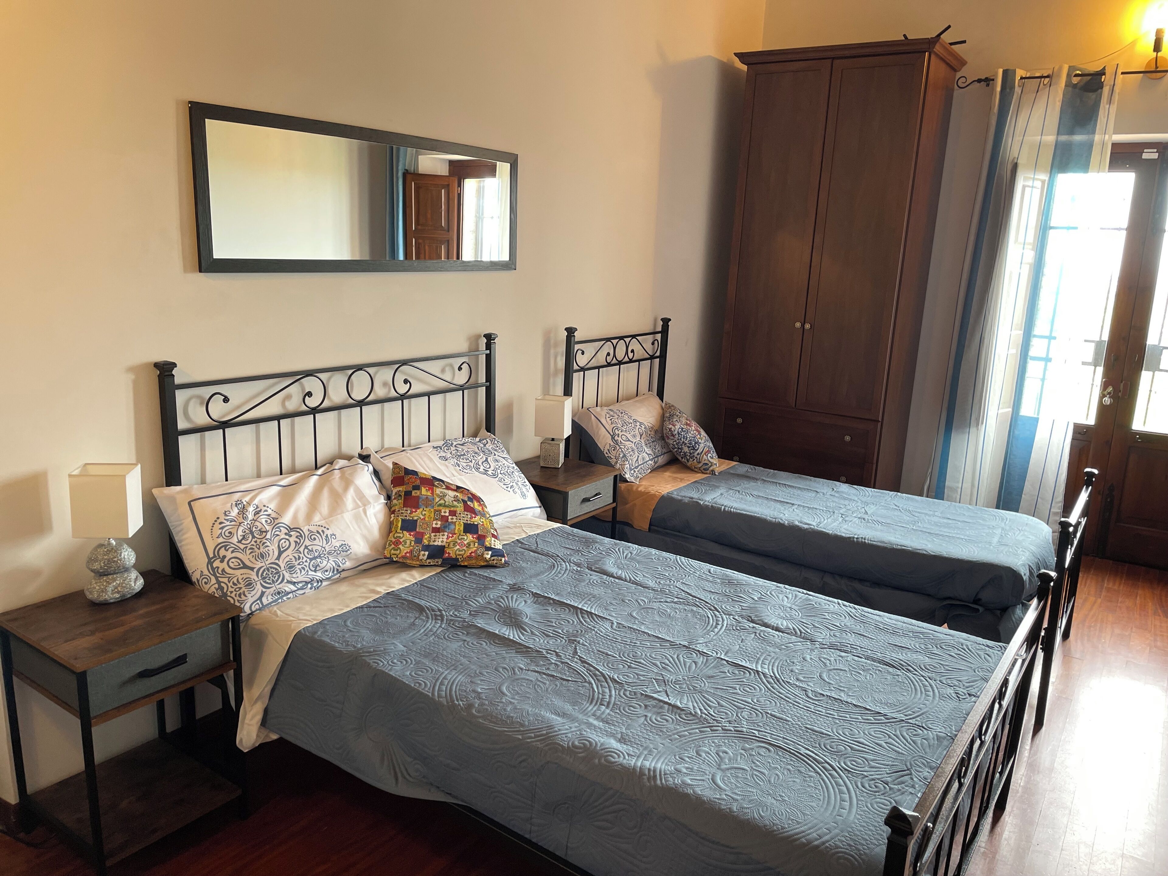 Cama doble con ropa de cama estampada, mesita de noche con lámpara, armario de madera y espejo en la pared.
