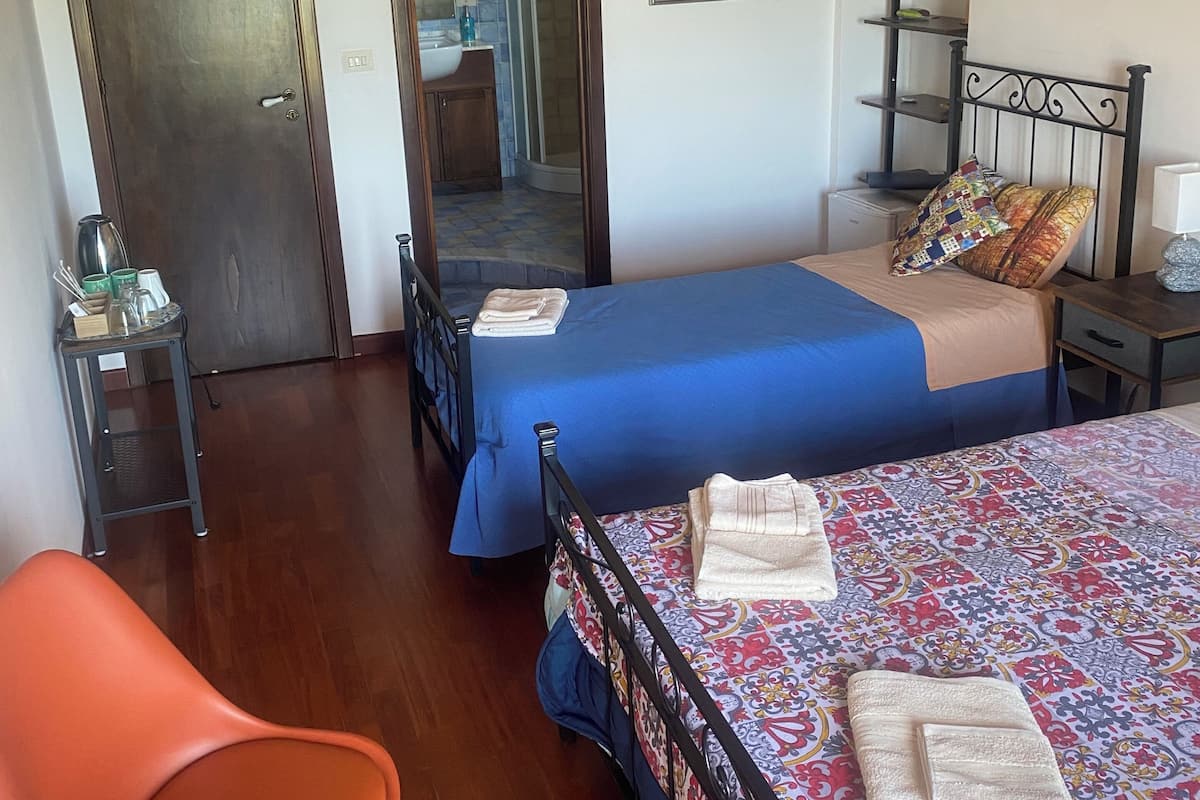 Un dormitorio con dos camas, una silla, una estantería y un baño visible a través de una puerta abierta.