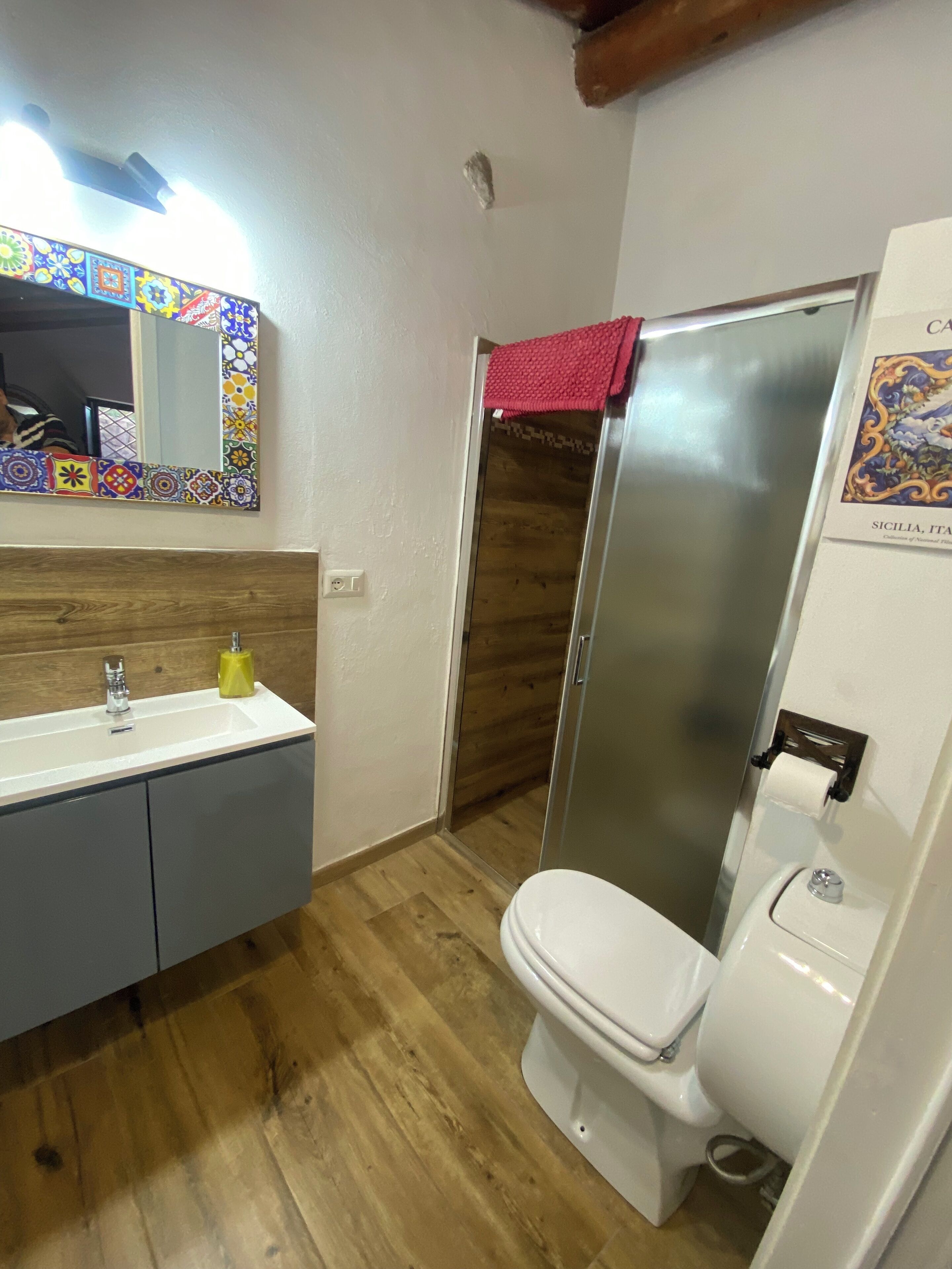 Uma casa de banho com chão de madeira, uma pia branca com dispensador de sabonete amarelo, uma sanita branca e um chuveiro com uma toalha vermelha.