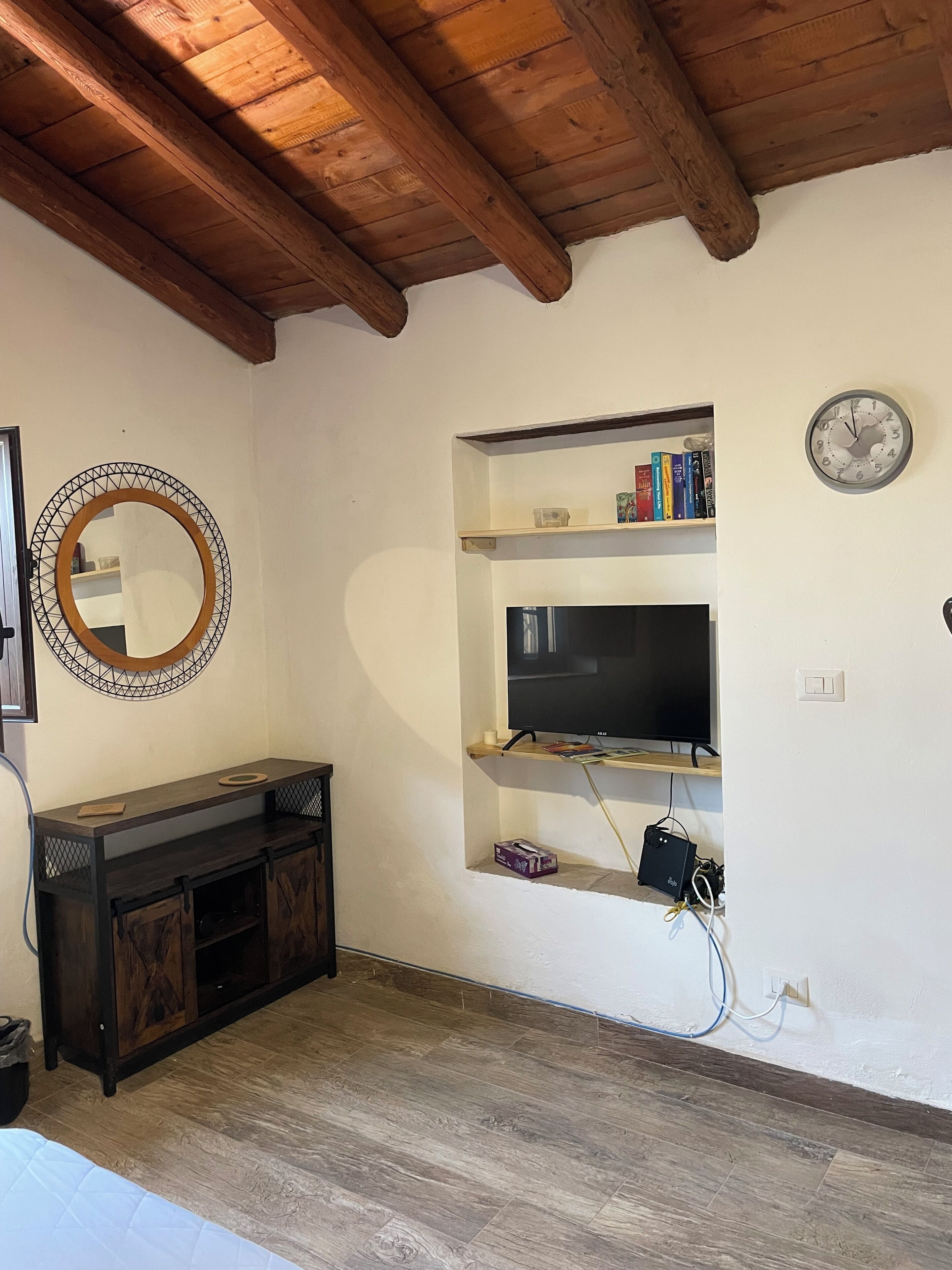 A living room with a wooden ceiling, a TV on a shelf, a clock, and a mirror on the wall.