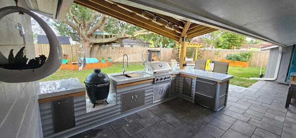Uma cozinha externa com churrasqueira embutida, pia e armários, sob uma pérgola de madeira.