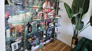 Une pièce avec une étagère vitrée exposant diverses bouteilles et verres, une grande plante en pot et un canapé vert.