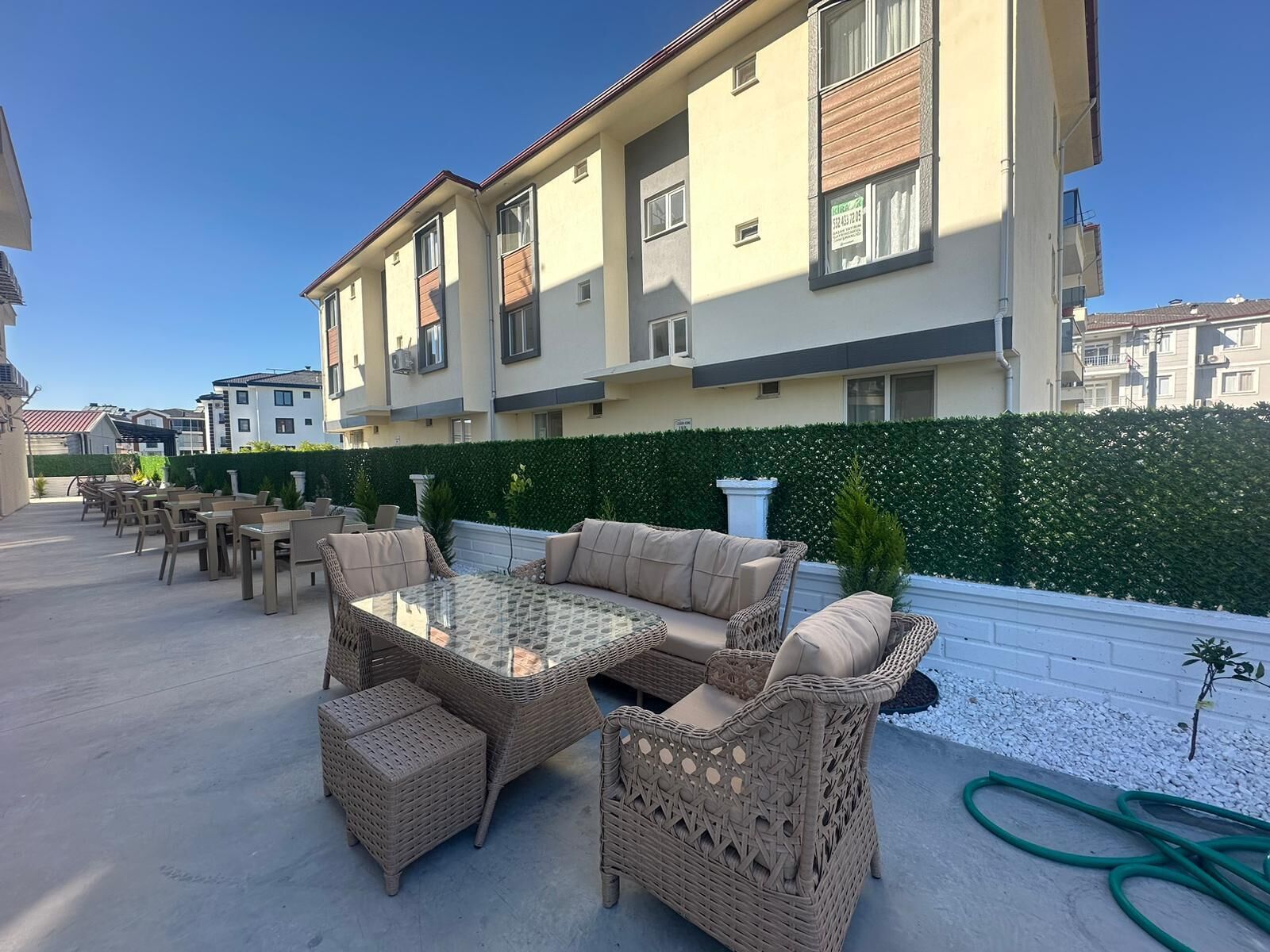 Un patio exterior con mobiliario de mimbre y una mesa con cubierta de vidrio, rodeado por un seto y una pared blanca.
