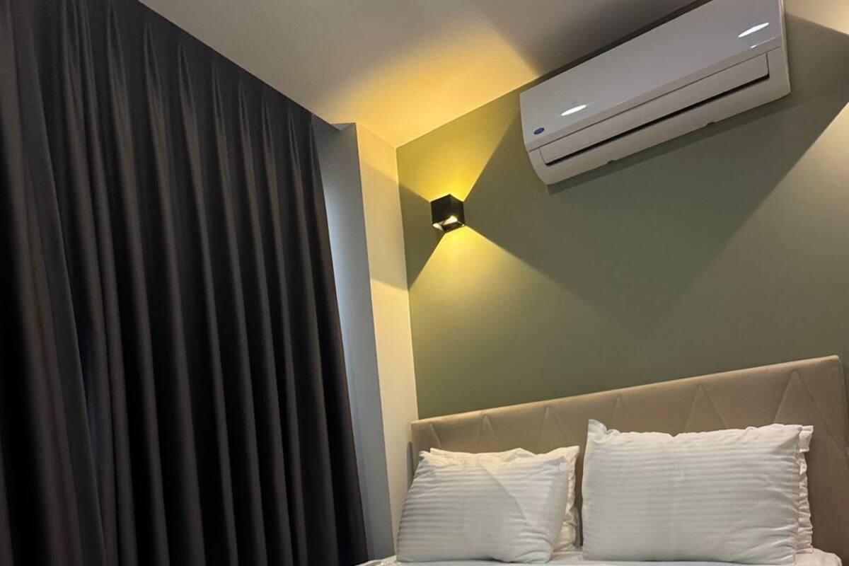 A hotel room with a neatly made bed, an air conditioning unit, a wall-mounted light, and a large window with curtains.