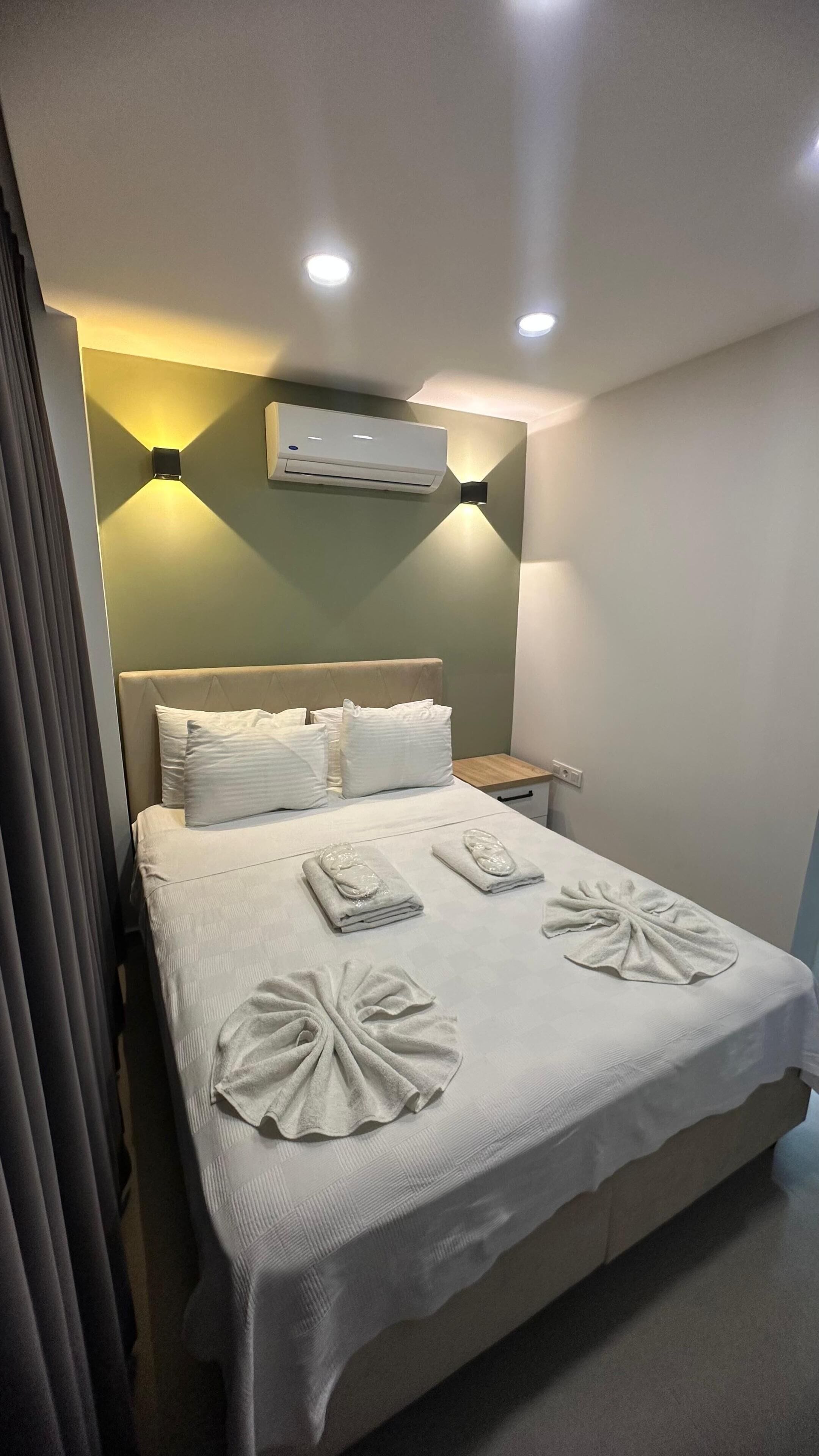 Habitación de hotel con cama, almohadas, mesita de noche, aire acondicionado y lámpara de pared.