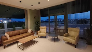 A modern living room with a sofa, two armchairs, and a coffee table. Large windows offer a view of the cityscape at dusk. - The Brothers Suite (Dalaman)