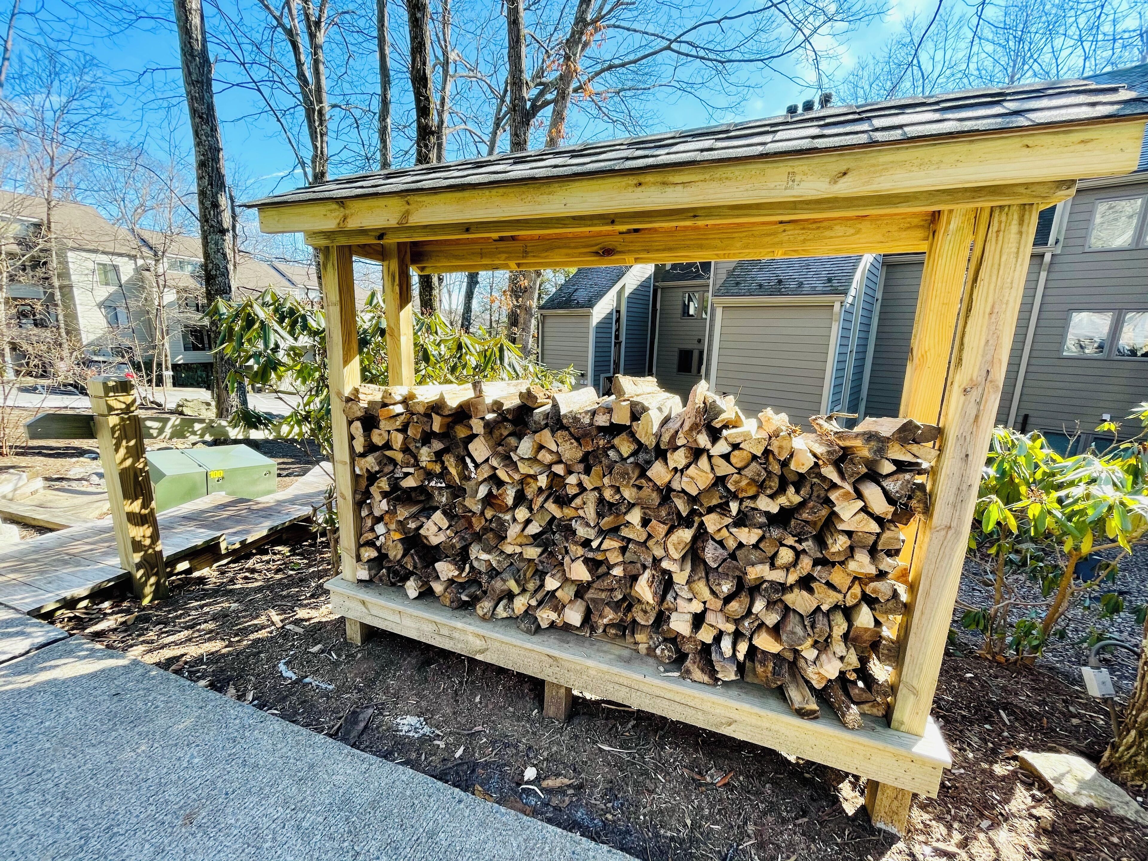 A wooden outdoor storage rack filled with firewood, surrounded by a garden and residential buildings.