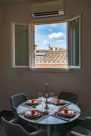 A dining area with a table set for four, a window with an open view of buildings, and an air conditioning unit above the window. - Giraldi House - 10 people (Firenze)