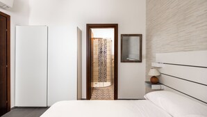 Ein modernes Hotelzimmer mit Bett, Nachttisch mit Lampe, Kleiderschrank und sichtbarer Dusche durch eine geöffnete Tür.