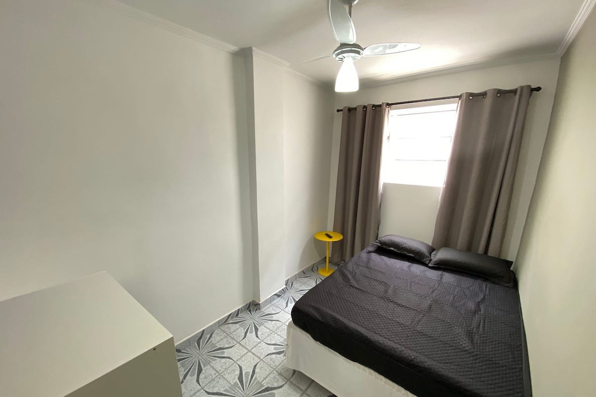 Une petite chambre à coucher, bien rangée, avec un lit, un ventilateur de plafond et une fenêtre avec des rideaux.