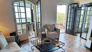 Un vaste espace de vie comprenant une table basse en verre, un canapé et un fauteuil. De grandes fenêtres et des portes-fenêtres offrent une vue sur l’extérieur.