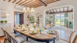 A dining area with a long table set for six, wooden beams on the ceiling, and a view of the outdoors through a glass door. - Fairytale Farmhouse Villa with Views and Pool (Lucca)