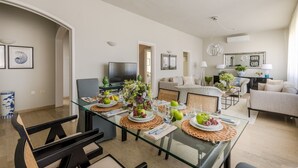A modern living room with a glass dining table set for four, a sofa, a TV, and a coffee table with plants. - Experience  Beachfront Coastal Elegance at Ocean Breeze (Lido di Camaiore)