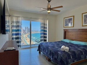 Un dormitorio con cama, escritorio y vistas a la playa y al paisaje urbano a través de ventanas amplias.