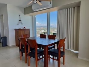Un comedor con una mesa y sillas de madera, vistas a un paisaje urbano a través de amplios ventanales y un equipo de aire acondicionado en el techo.