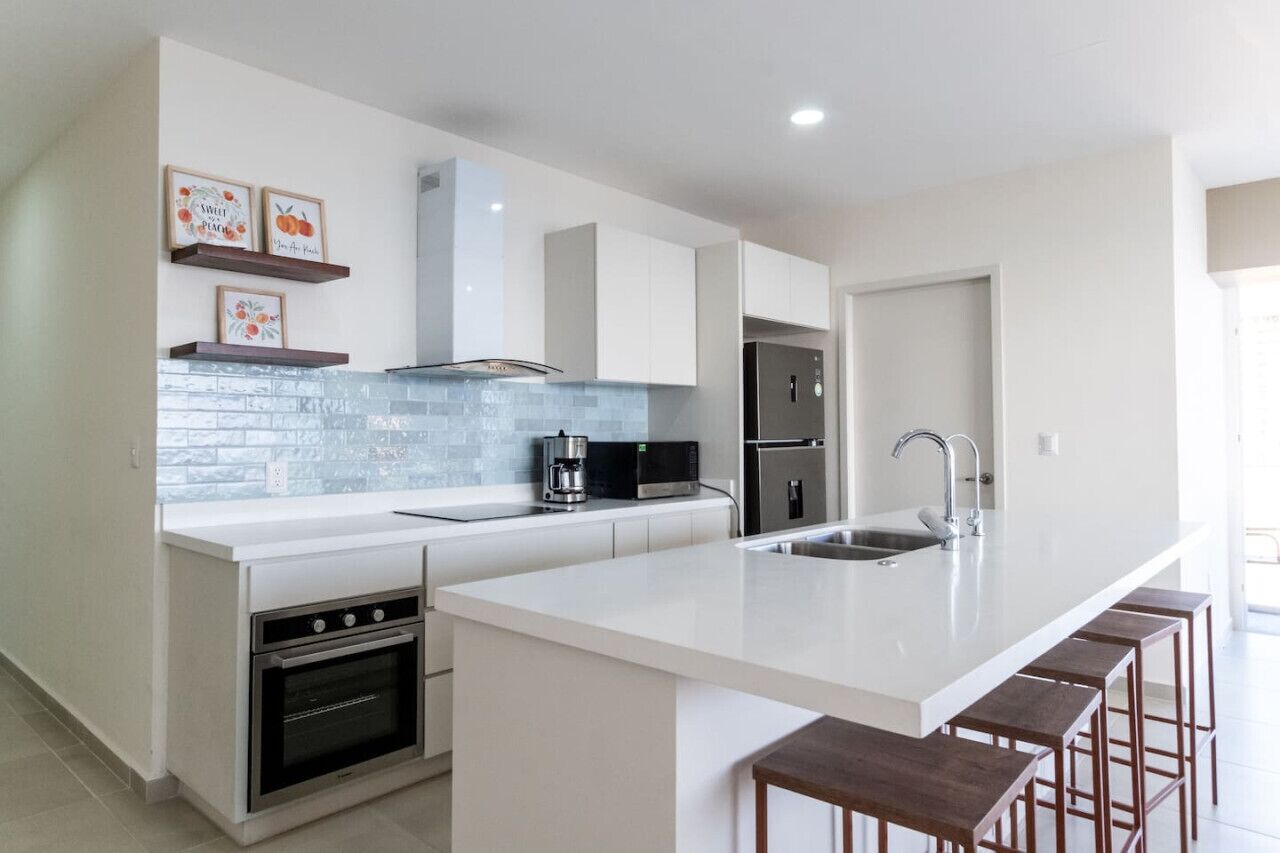 Una cocina moderna con armarios blancos, una isla grande con taburetes de bar, horno empotrado, microondas y un refrigerador de acero inoxidable.